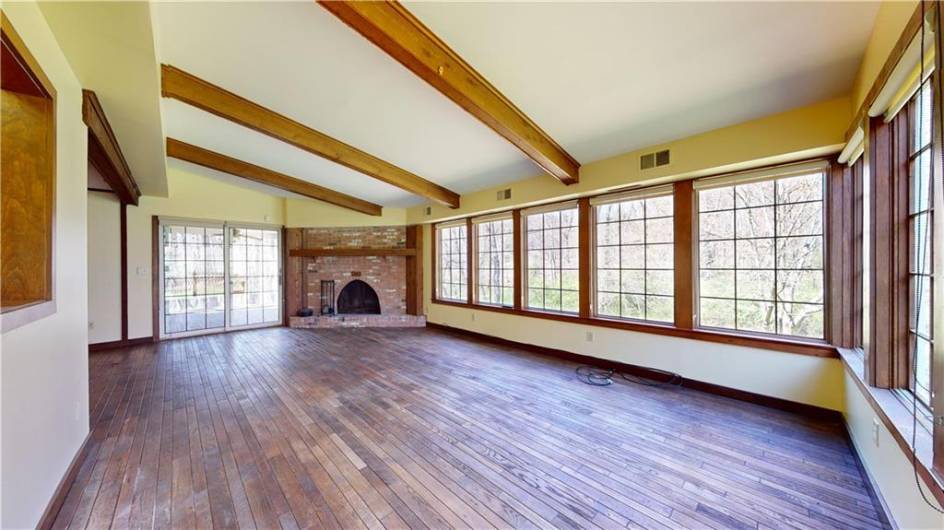 The family room features a vaulted ceiling adorned with exposed wooden beams that complement the rich, dark wood floor. A series of large windows offer a panoramic view of the serene woods outside and flood the room with natural light.