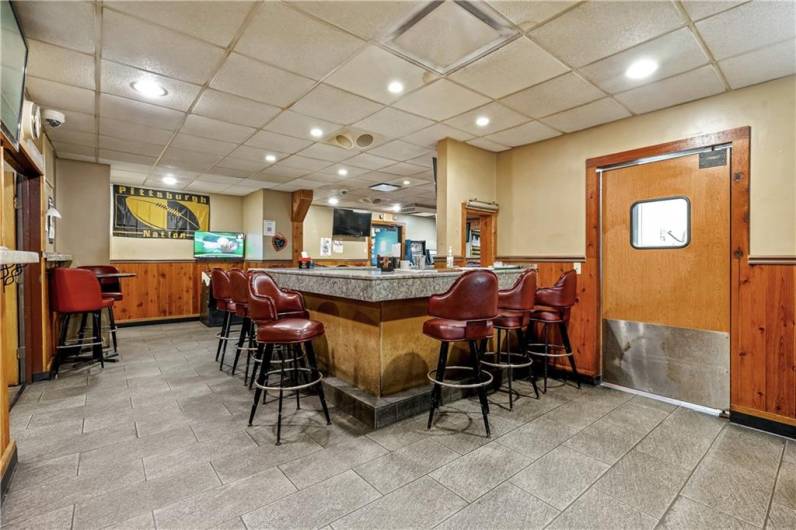 Corner of bar area with door to kitchen on right side and entrance to dining area on left wall in photo