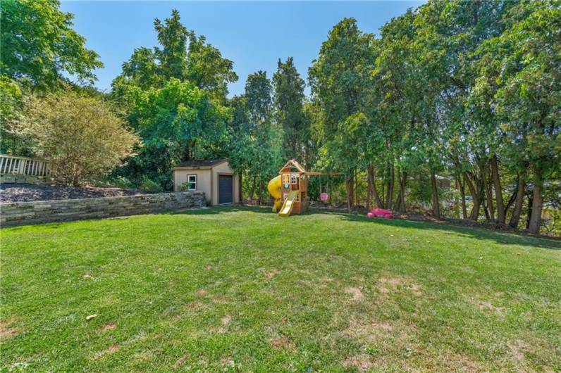 Park like tree lined level back yard with storage shed and playset.