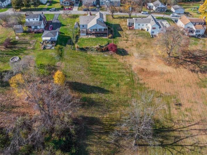 This aerial shot captures the spacious backyard of the property, showcasing neighboring homes and a bird’s eye view of the layout. A fire pit area is visible on the left side, and the fenced yard provides a clear view of property boundaries. The neighboring yards and tree lines frame the area, creating a sense of community and open space.