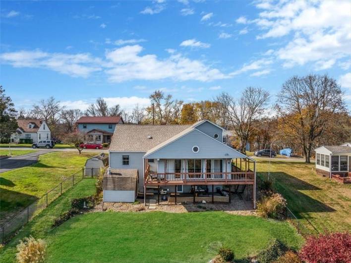 A closer perspective of the deck area reveals more details, including access stairs, storage space underneath, and landscaping that frames the property. The siding and roofing details of the house are also clearly visible.