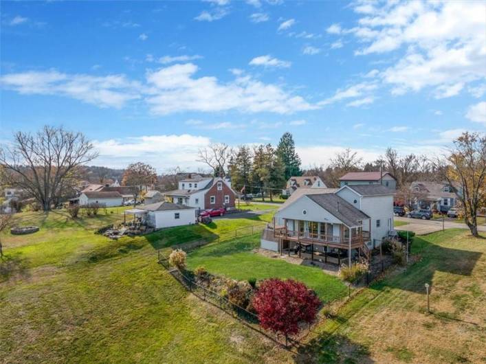 This shot provides a clear view of the entire home, backyard, and neighboring structures. The fenced yard is well-defined, and you can see landscaping elements like shrubs, small trees, and open green spaces.