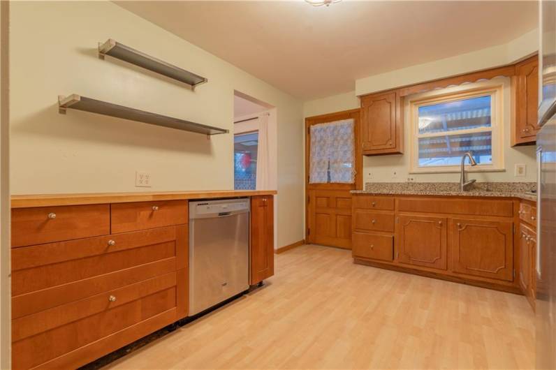 A view of the left side of the kitchen with butcher block counters and stainless dishwasher and open shelves. The back door allows access to the covered rear patio.