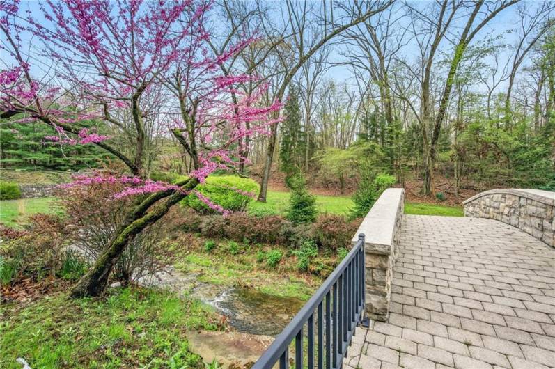 Charming stone bridge crosses a gentle stream, all framed by vibrant spring blossoms and lush greenery.