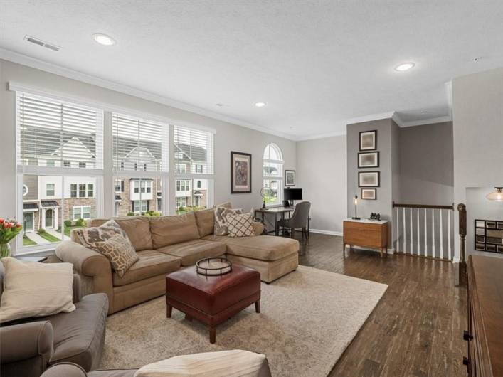 stunning great room offers a perfect blend of style, space, and functionality. Bathed in natural light from a wall of oversized windows, the open layout features rich hardwood flooring and recessed lighting for a bright, welcoming atmosphere.
