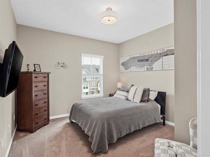 This comfortable secondary bedroom offers a cozy and inviting atmosphere with neutral-toned walls and plush carpeting. A large window allows natural light to brighten the space, while the room's layout easily accommodates a full-sized bed and additional furniture. Stylish yet functional, this room is perfect for guests, family members, or a home office. Thoughtful details, including a wall-mounted TV and tasteful decor, complete the look of this well-appointed space.