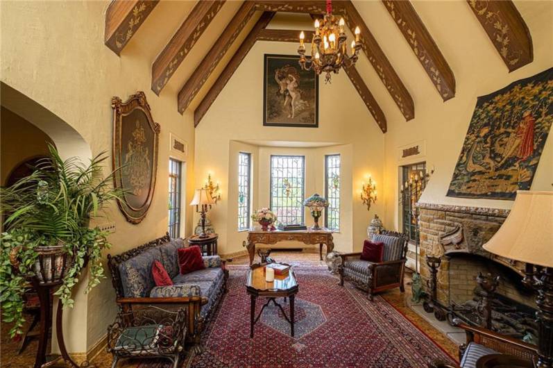 A stately living room complete with African Cyprus beams and an amazing stone fireplace that has been converted to gas for ease of use. Also, note the 3 Musketeers stained glass windows at the curve in the room.