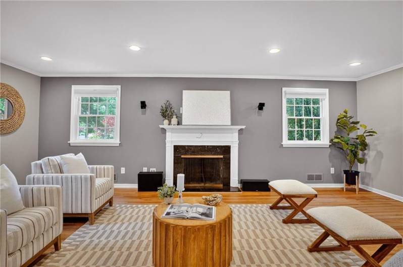 A white fireplace serves as the focal point, flanked by large windows that fill the room with natural light. Recessed lighting and wood floors enhance the bright, welcoming ambiance.