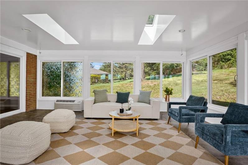 A bright sunroom with floor-to-ceiling windows and skylights, featuring stone-look tile flooring, white trim, and a cozy, light-filled atmosphere ideal for relaxing or entertaining year-round.