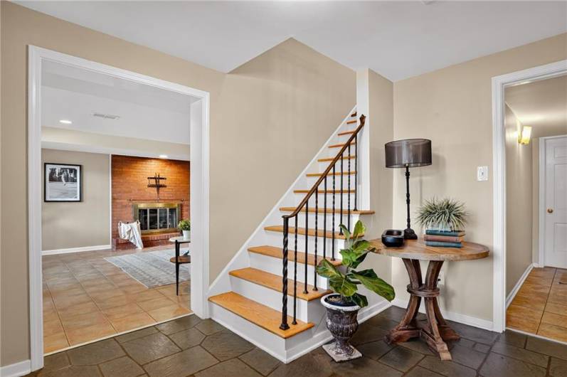 A welcoming foyer with slate tile flooring, a wooden staircase featuring iron spindles. The view leads into a cozy family room with a brick fireplace.