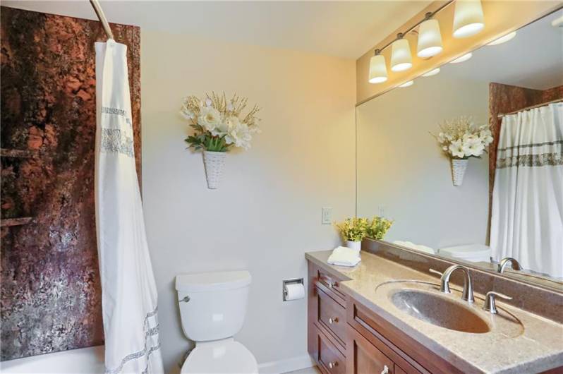 This stylish full bath features a rich wood vanity with solid-surface counters, a large framed mirror, and elegant lighting. The striking shower surround adds a touch of drama and sophistication to this thoughtfully designed space.