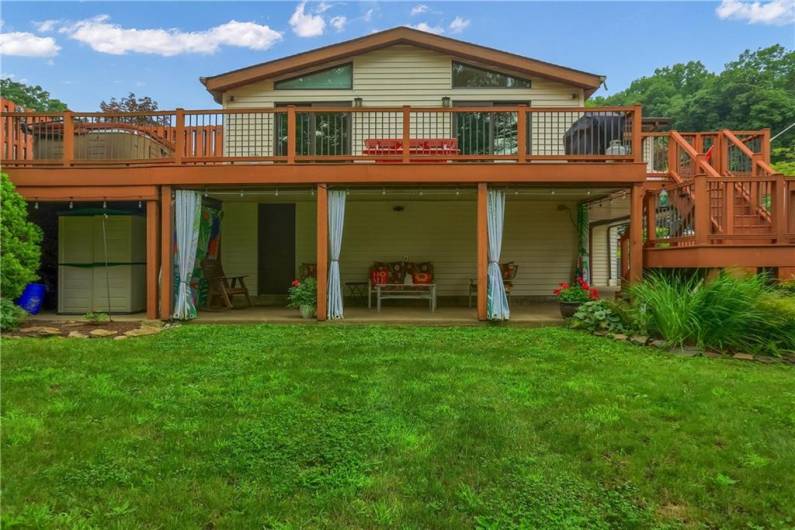 The back of the home is an entertainer’s dream, featuring a spacious upper deck with modern railings and dual access points, plus a covered patio below with underdecking and vibrant outdoor curtains for added privacy and charm. Surrounded by lush greenery, it offers the perfect blend of relaxation and outdoor living.